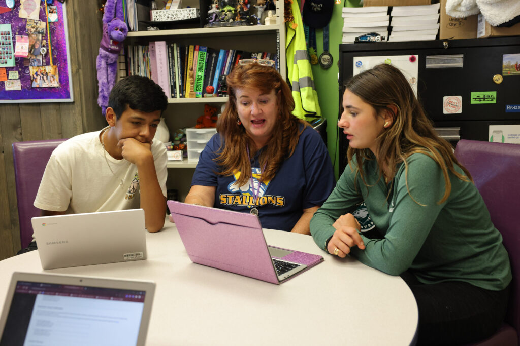 Looking at a laptop, two students sit on either side of a teacher at a round table in a classroom with a purple bulletin board, a bookshelf, and a filing cabinet behind them.