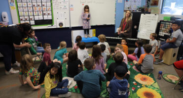 A class of kindergarten students at SCVi is sitting on a rug in front of a whiteboard. Once student is standing up in front of the class.
