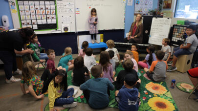 A class of kindergarten students at SCVi is sitting on a rug in front of a whiteboard. Once student is standing up in front of the class.