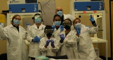 Group of SCVi students and teachers pose in front of a low gray table in white lab coats, safety goggles and masks. A couple students hold vials. Two machines reading “Air Science” tower in the background