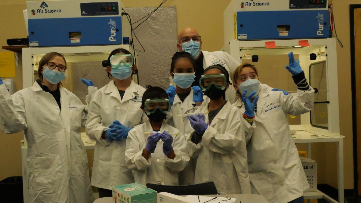 Group of SCVi students and teachers pose in front of a low gray table in white lab coats, safety goggles and masks. A couple students hold vials. Two machines reading “Air Science” tower in the background