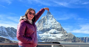 Santa Clarita Valley International (SCVi) School alum Emma Hild stands in front of a mountain with her arm raised making it look like she’s touching the peak.