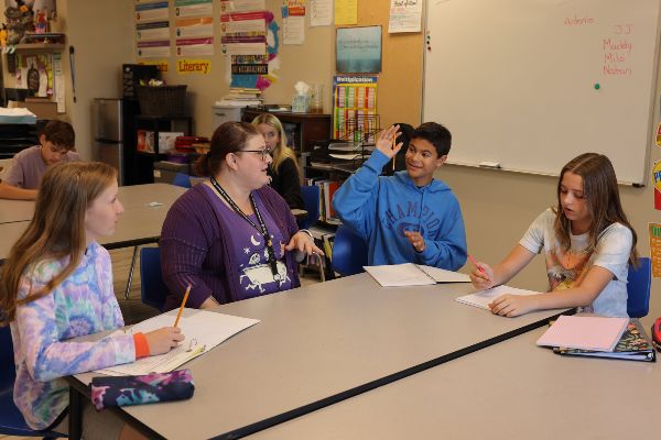 Three Santa Clarita Valley International (SCVi) students sit with an adult at a classroom table in a discussion. They have binders with paper on the table and are using pencils. One student is raising his hand.