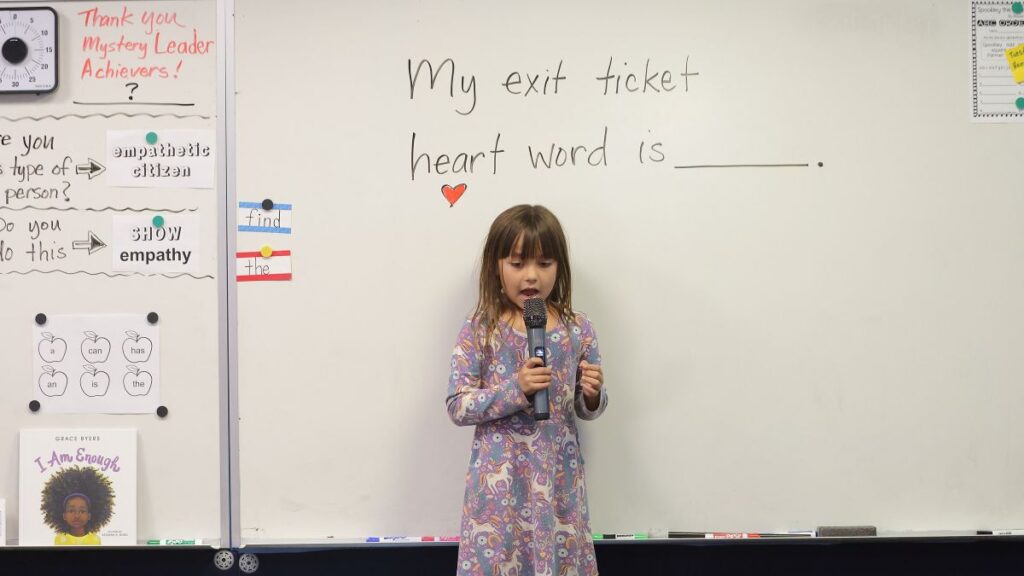 Santa Clarita Valley International (SCVi) School student stands in front of a classroom white board that says “My exit ticket heart word is” with a microphone in her hand.