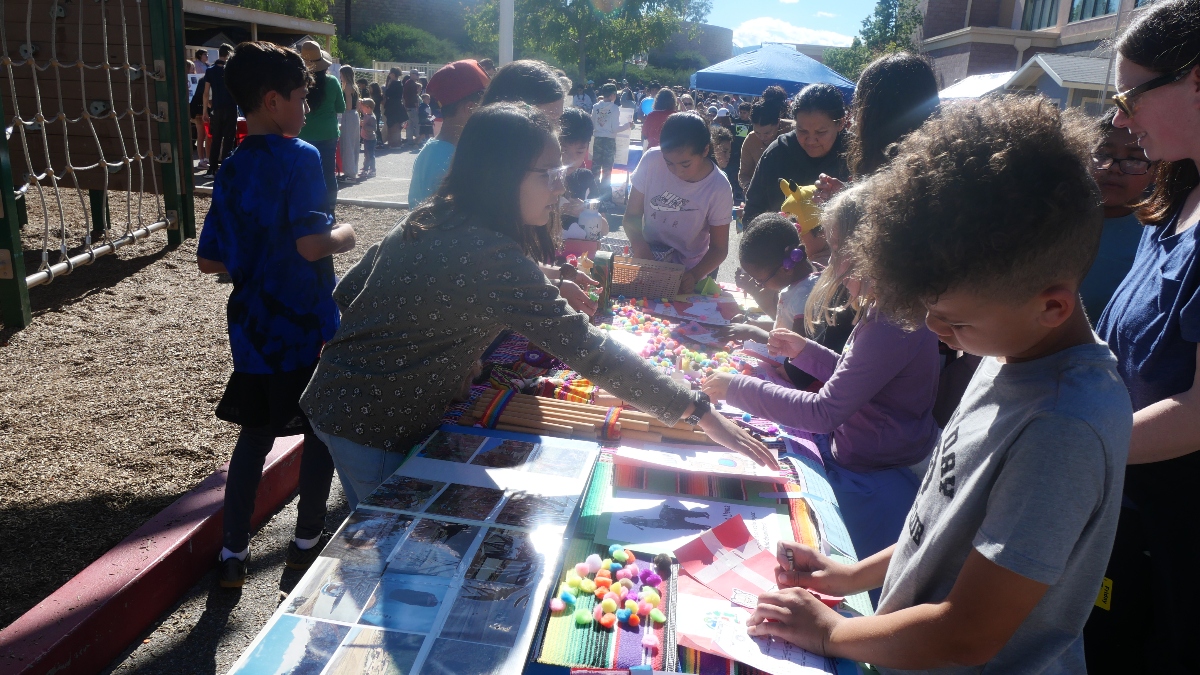 Santa Clarita Valley International (SCVi) students surround a long table outside covered with photos and colorful drawings. Students color and one is reaching across the table.