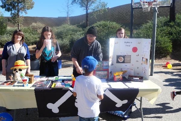 Santa Clarita Valley International (SCVi) Charter School students stand outside at a long table with a “Japanese Foods” labeled trifold board and small boxes in front of it. One student in glasses is smiling down at a younger student in a blue baseball cap who has their back to the camera.