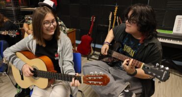 Santa Clarita Valley International (SCVi) students sit playing guitar -one acoustic and one electric), both smiling and looking off-camera. More students practice in the background.