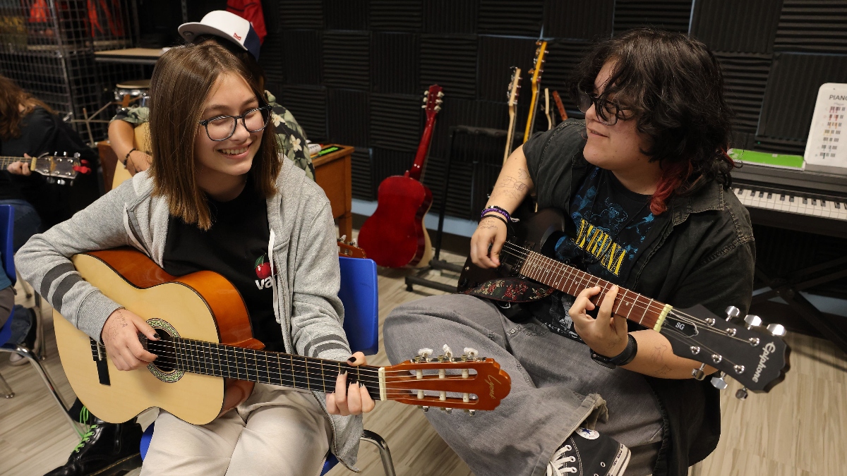 Santa Clarita Valley International (SCVi) students sit playing guitar -one acoustic and one electric), both smiling and looking off-camera. More students practice in the background.