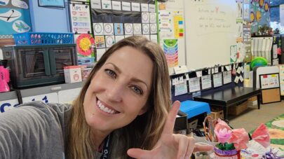 Santa Clarita Valley International (SCVi) teacher Amy Ciceri smiles looking at the camera with her colorful classroom with many arts and crafts behind her.