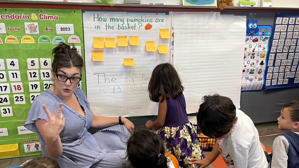 A group of Santa Clarita Valley International (SCVi) students sit on a rug around a teacher who is also sitting and holding up two fingers on her hand while another student writes something on a big pad of paper on the wall that says, “How many pumpkins are in the basket?”.