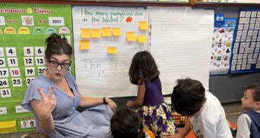 A group of Santa Clarita Valley International (SCVi) students sit on a rug around a teacher who is also sitting and holding up two fingers on her hand while another student writes something on a big pad of paper on the wall that says, “How many pumpkins are in the basket?”.