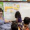 A group of Santa Clarita Valley International (SCVi) students sit on a rug around a teacher who is also sitting and holding up two fingers on her hand while another student writes something on a big pad of paper on the wall that says, “How many pumpkins are in the basket?”.