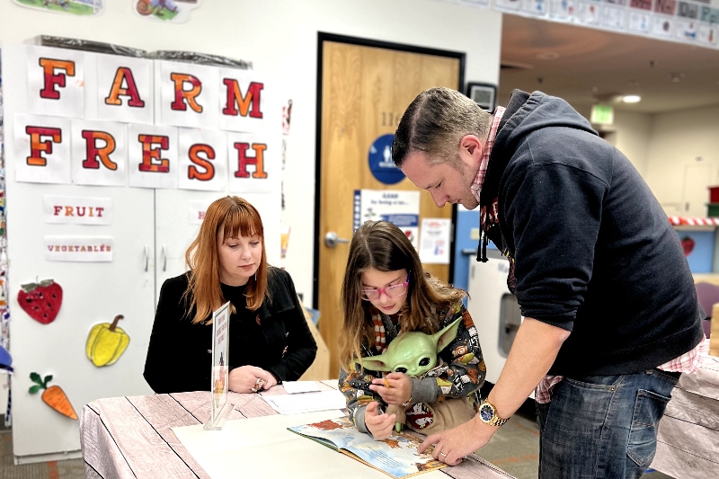 A Santa Clarita Valley International (SCVi) family looks at a book on a table together inside a colorful classroom with “FARM FRESH” letters on the wall and a strawberry, yellow pepper, and carrot picture underneath. 