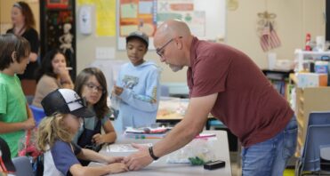 Santa Clarita Valley International (SCVi) teacher reaches across the table to help a student. Other students mill around in the background, waiting for project materials.