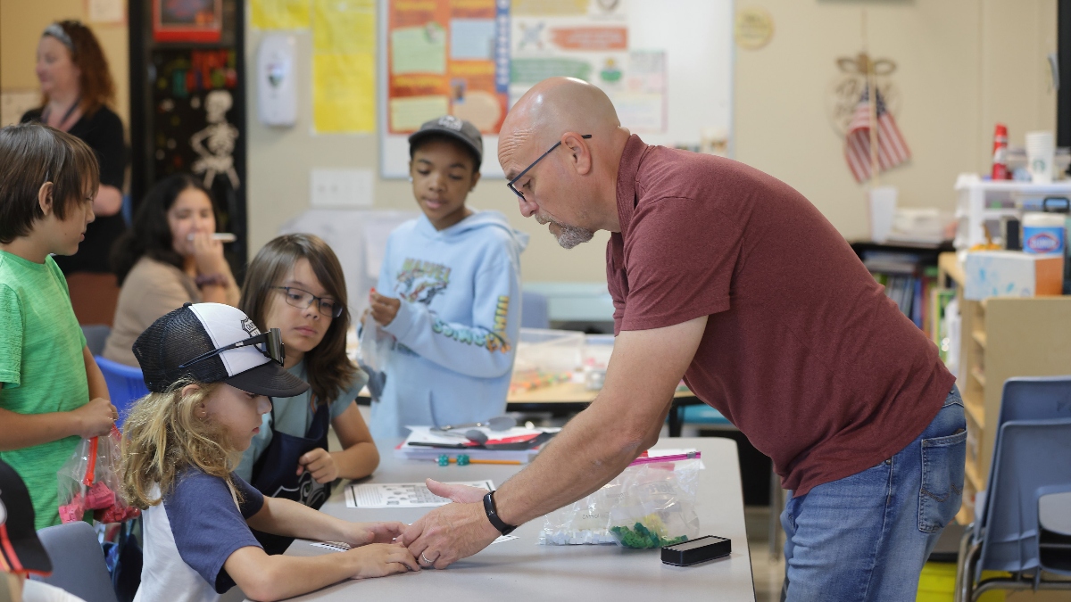 Santa Clarita Valley International (SCVi) teacher reaches across the table to help a student. Other students mill around in the background, waiting for project materials.