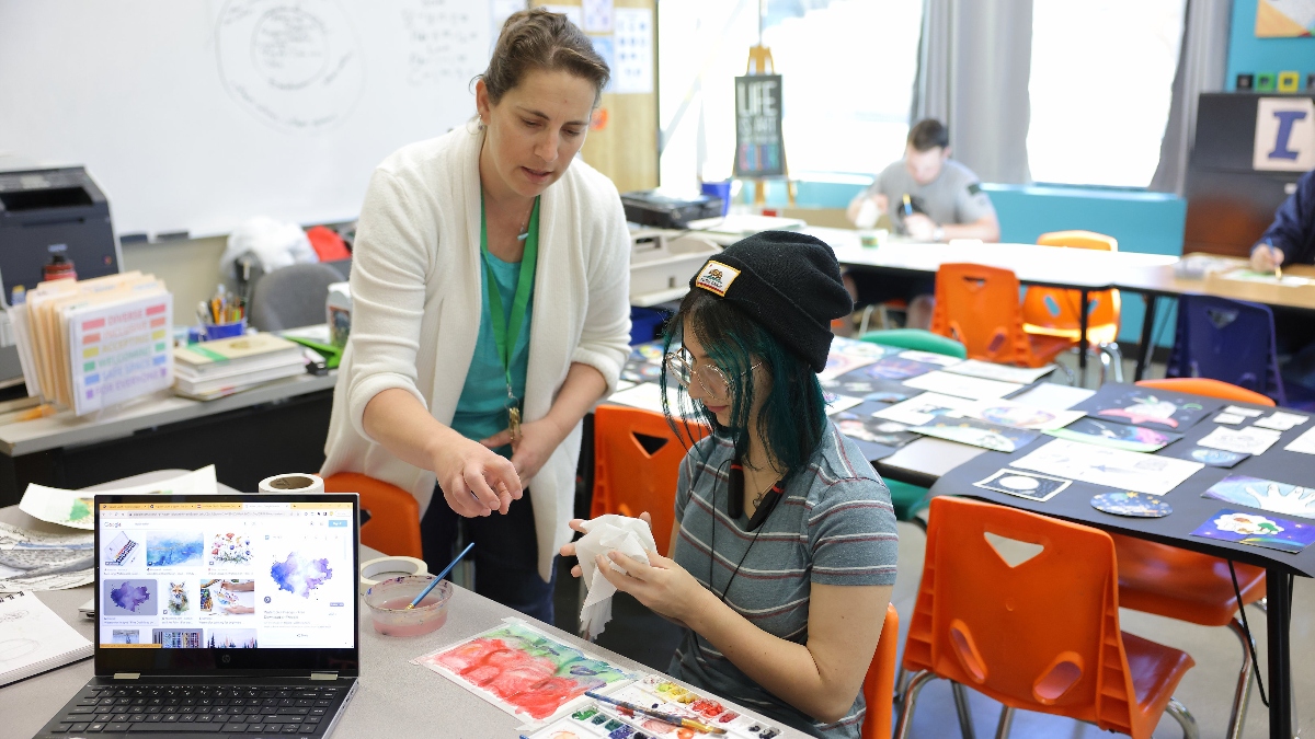 A Santa Clarita Valley International (SCVi) teacher extends her arm over a student’s artwork while the student holds a scrunched paper towel above a piece of paper with painted colors on it at a classroom table. There’s also a laptop on the table and another table in back of the student with paintings and drawings laid out on it.